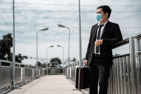 Unemployed stressed young Asian business man wear mask carry a job bag looking for work in suit covering face with Leaning against a bridge rail. Failure and layoff concept to the disease Covid-19の写真素材