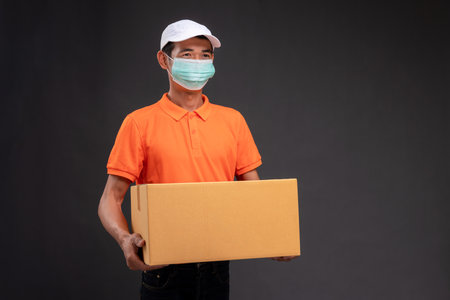Delivery asian man or courier wearing an orange masked uniform holds a cardboard box in the coronavirus concept studio.の写真素材