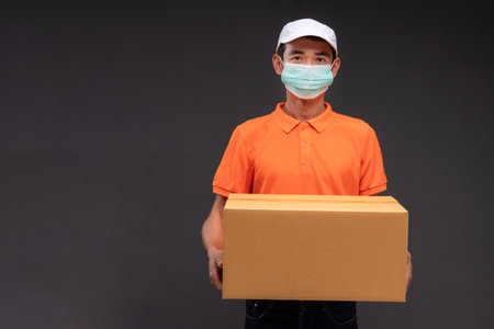 Delivery asian man or courier wearing an orange masked uniform holds a cardboard box in the coronavirus concept studio.の写真素材