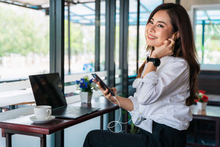 Happy asian beautiful woman working on a laptop and listen to music on your smartphone with headphones sitting in the coffee shop.の写真素材