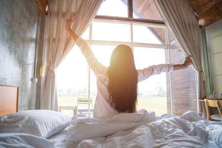 Beautiful asian women is waking up in the morning, Stretches in the Bed, Sun Shines on Her From the Big Window.の写真素材
