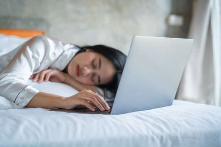 Beautiful asian girl in white pajamas sleeps at bed with laptop, laptop after work.の写真素材