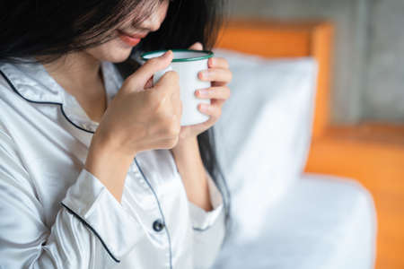 Closeup hand of pretty young woman sitting on the bed and enjoying first morning coffee on sunshine.の写真素材