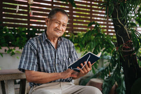 Happy senior thai man sitting on marble chair under the tree and reading book, Healthy senior conceptの写真素材
