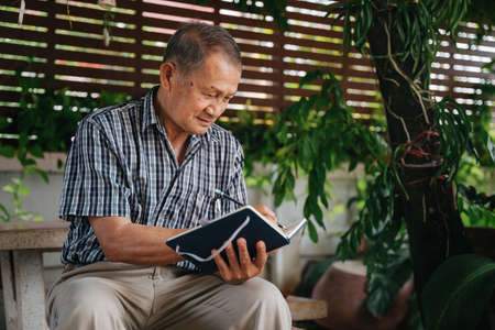 Senior thai man sitting on marble chair under the tree and write notes, Alzheimer's disease concept.