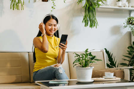 Portrait of happy asian woman chat with mobile phone and listen to music with headphones in coffee shop, cafeの写真素材