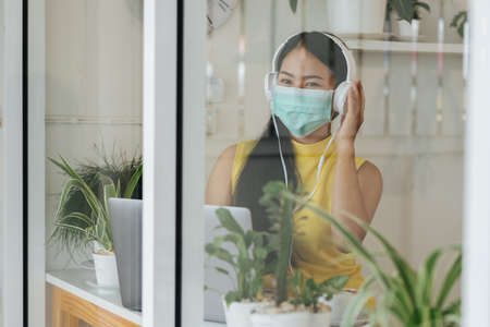 Portrait of happy asian woman wear a mask to prevent the spread of coronavirus or COVID-19 and listen to music with headphones in coffee shop, cafeの写真素材