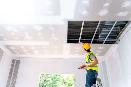 Asian construction worker assemble a suspended ceiling with drywall and fixing the drywall to the ceiling metal frame with screwdriver. Renovation, construction and ceiling installation concept.の写真素材