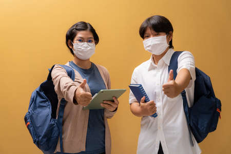 Two girls giving wear a protective mask with thumb up and learning equipment in student uniform, Education concept and prevention of the spread of corona virus.の写真素材