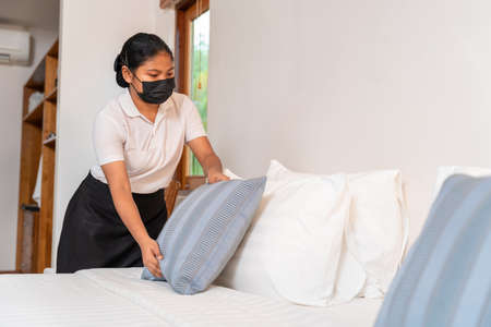 Asian maid setting up pillow on bed sheet in hotel room.