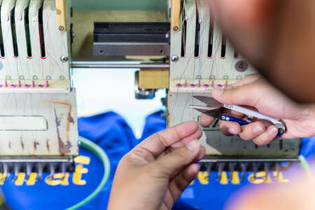 A worker use scissors to cut yarn on sewing machine. Embroidery area in textile factory in industrial zone with modern machinery and technology systems.の写真素材