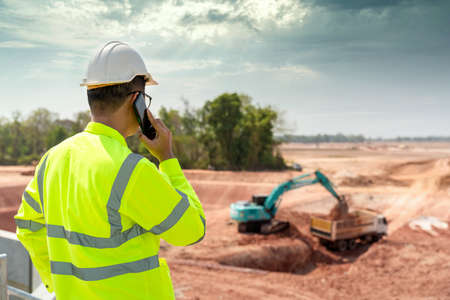 A civil engineer is using a construction telephone on the construction site in the background with loaders and dumpers.の写真素材