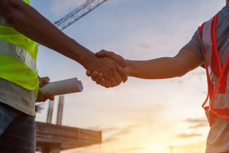 Hand of architect civil and engineer in protective helmets and vests are shaking hands while working in construction site.の写真素材