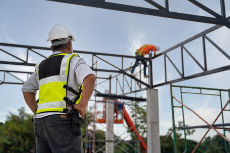 Contractor stood to watch the progress of welder is welding steel on a steel roof truss. Fall arrestor device for worker with hooks for safety body harness. Worker in construction site.の写真素材