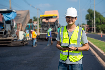 Asian handsome civil engineer under inspection and survey workplace by tablet with Asphalt paver & road roller & dump truck and worker construction road background.の写真素材