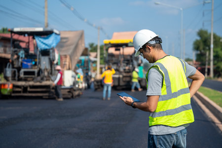 Asian civil engineer working on a road construction site with Asphalt paver & road roller & dump truck and worker construction road background.の写真素材
