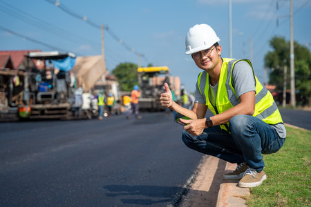 Asian young civil engineer showing confidence with thumb raised at road construction site with Asphalt paver & road roller & dump truck and worker construction road background.の写真素材