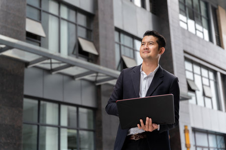 Handsome Asian business man holding laptop looking away happily in urban public space.の写真素材