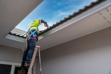 Asian roofer wearing safety harness belt using air or pneumatic nail gun and installing house roof tiles on top new roof at home under construction. Roofing tools.の写真素材
