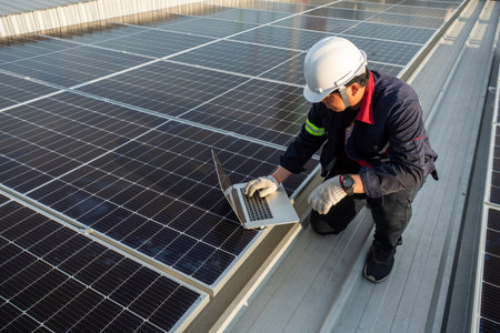 Asian engineer using laptop inspecting solar photovoltaic panels on the roof of an industrial...