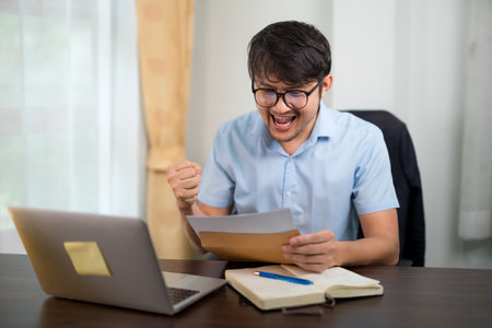A handsome young man in glasses succeeds while reading acceptance letter with a laptop on his desk at home.の写真素材
