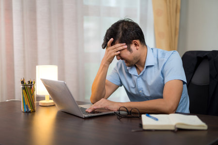 Asian man having severe migraine headache due to long hours of work and reserving a laptop at his desk at home.の写真素材