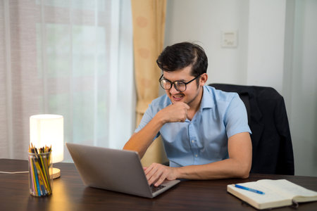 Asian handsome man in glasses working on laptop with smile at home desk.の写真素材