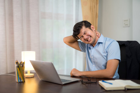 Asian young man with neck pain or office syndrome after working long hours with a laptop on your desk at home.の写真素材
