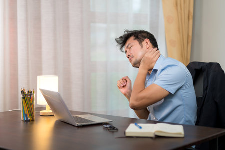 A man with neck pain Or office syndrome is doing self-massage of the neck on a desk with a laptop at home.の写真素材