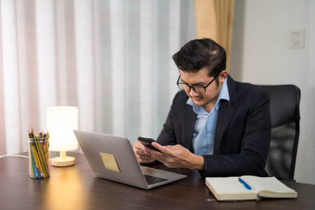 Asian businessman is using smartphone to check messages on desk with laptop at home.の写真素材