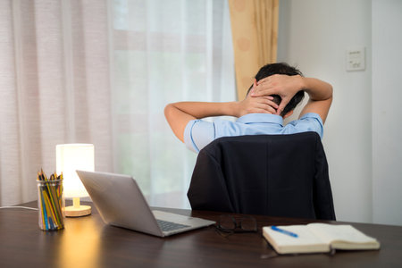 Asian young man with neck pain Or office syndrome is doing self-massage of the neck on a desk with a laptop at home.の写真素材