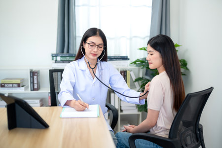 Asian female doctor using stethoscope to check patient's heartbeat with note taking at hospital. Medicine. Elderly health. Healthcare and medical concept.の写真素材