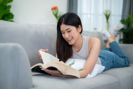 Asian woman lying and reading a book on the sofa in the living room in the morning.の写真素材