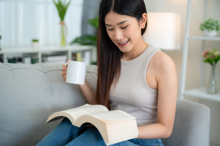 Close up of Asian young woman reading a book with coffee on the sofa in the living room in the morning.の写真素材