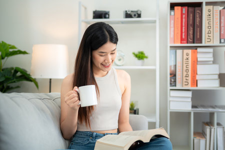 Asian young woman holding a coffee cup together reading a book on the sofa in the living room.の写真素材