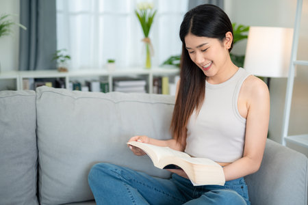 Asian young woman sitting happily reading a book on the sofa in the living room. brain food ideas.の写真素材