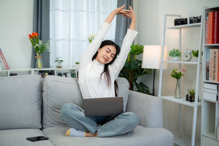 Asian woman stretching out her arms to relax on the sofa in the living room after working for a long time on the laptop.の写真素材