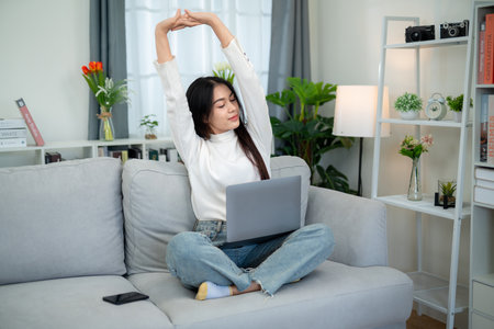 Asian woman stretching on the sofa in the living room after working for a long time on the laptop.の写真素材