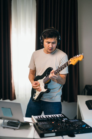 Asian male composer creating a track with guitar and MIDI piano, a cozy home studio filled with inspiration and soft ambient light.の写真素材