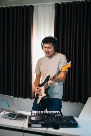 Asian male blending guitar melodies with electronic beats, surrounded by modern audio equipment and a digital workstation in a stylish home setupの写真素材