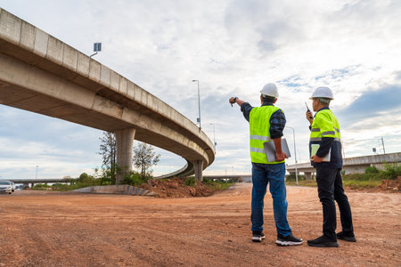 Two construction engineers stand on a muddy worksite, pointing and communicating via radio under a massive curved highway overpass.の写真素材