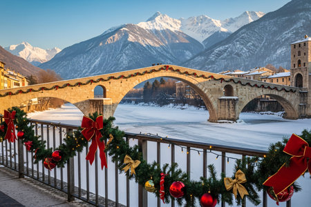 Historic stone bridge adorned with festive Christmas garlands, bows, and lights over a frozen river with snowy mountains in the background at sunsetの素材