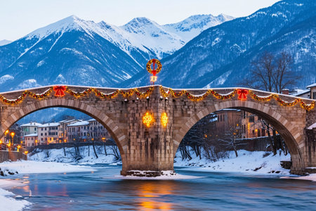 Historic stone bridge adorned with festive Christmas garlands, bows, and lights over a frozen river with snowy mountains in the background at sunsetの素材