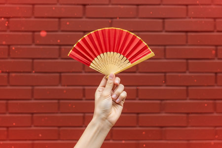 Chinese folding fan held by a hand is prominently displayed against the textured surface of a vibrant red brick wall.の素材