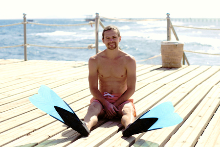 A young handsome man is sitting  in flippers on the bridge near blue seaの写真素材