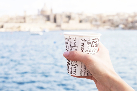 A female hand is holding a cup of coffee in front of sea and beautiful view on Valletta,Maltaの写真素材