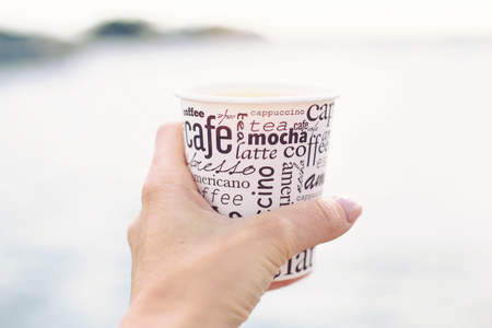 A female hand is holding a cup of coffee in front of sea and beautiful viewの写真素材