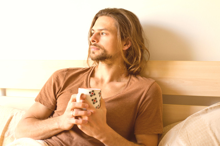 a young serious man  is looking out of the window with a cup of tea in his hands thinking about something lying in bed after wake upの写真素材