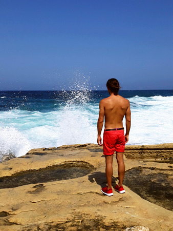 handsome young tanned man in shorts, stand on the edge of rock with back looking at sea waves, blue sky,calm down, outdoor sexy fashion model,believe in hope,traveling tourist,feel freedom,relax,independenceの写真素材