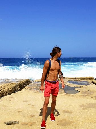 handsome young tanned man in shorts,with backpack, look back at sea waves, blue sky,calm down, outdoor sexy fashion model,traveling tourist,feel freedomの写真素材